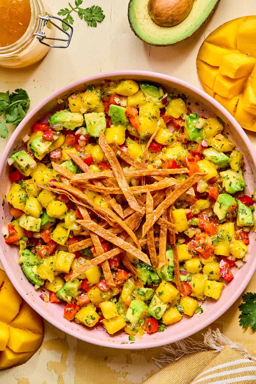 A fresh mango avocado salad with diced mango, avocado, tomato, and herbs, topped with crispy tortilla strips in a pink bowl. Avocado halves, mango slices, and a jar of dressing surround the bowl for a fresh presentation.