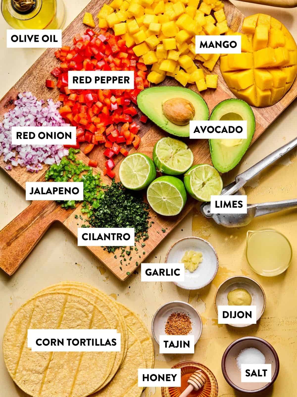 A vibrant mango avocado salad spread featuring diced mango, red pepper, onion, jalapeno, cilantro, avocado, limes, olive oil, Dijon mustard, Tajín, honey, salt and corn tortillas arranged on a wooden board over a yellow surface.