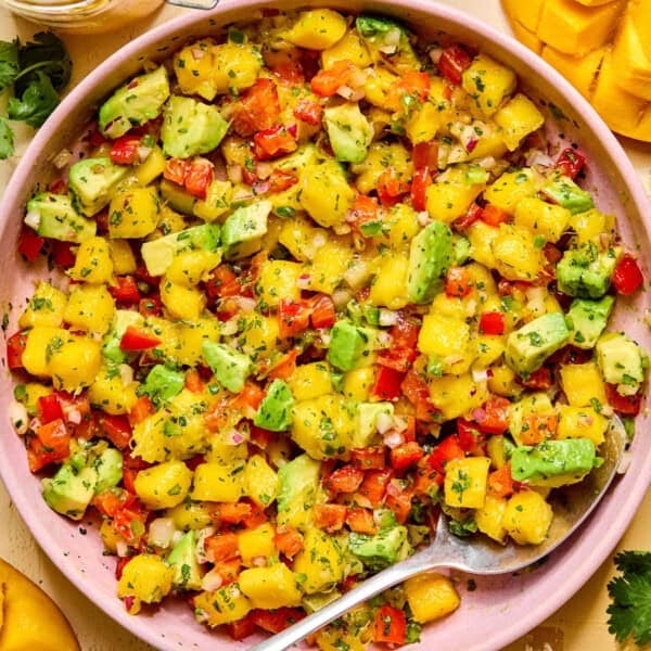 A bowl of colorful mango avocado salad features diced mango, avocado, red bell pepper, onion, and herbs, all mixed together with a spoon for a fresh and vibrant salsa.