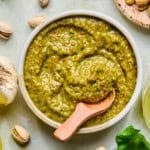 A bowl of green pistachio pesto sauce with a wooden spoon, surrounded by pistachios, a garlic bulb, and fresh basil leaves on a light surface.