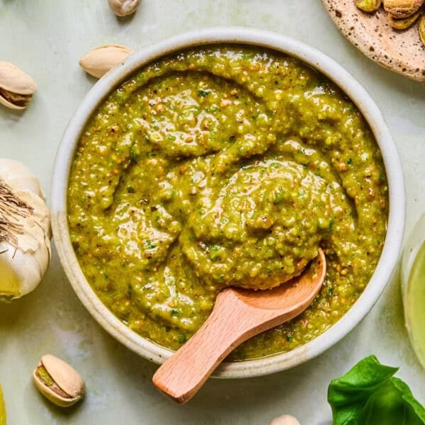 A bowl of green pistachio pesto sauce with a wooden spoon, surrounded by pistachios, a garlic bulb, and fresh basil leaves on a light surface.