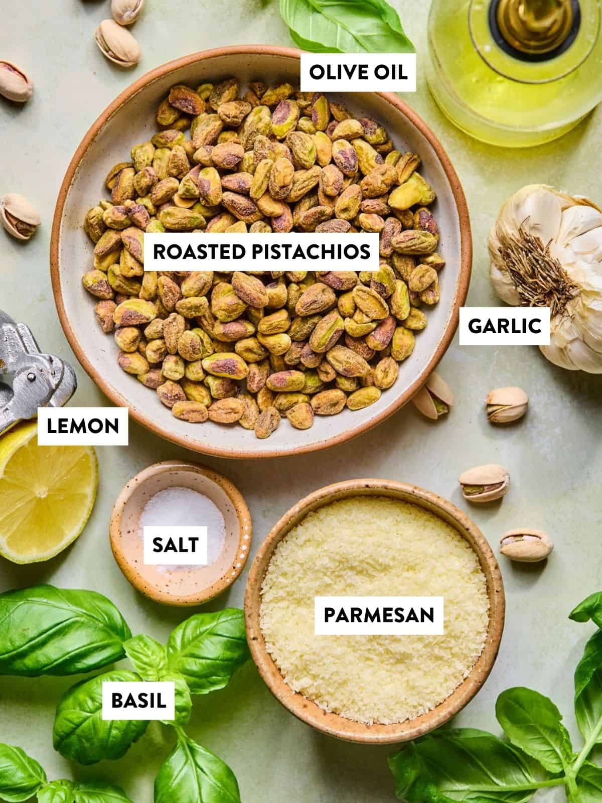 A flat lay of ingredients for pistachio pesto: a bowl of roasted pistachios, grated parmesan, fresh basil leaves, a halved lemon, garlic bulb, salt, and olive oil—all beautifully arranged and labeled.