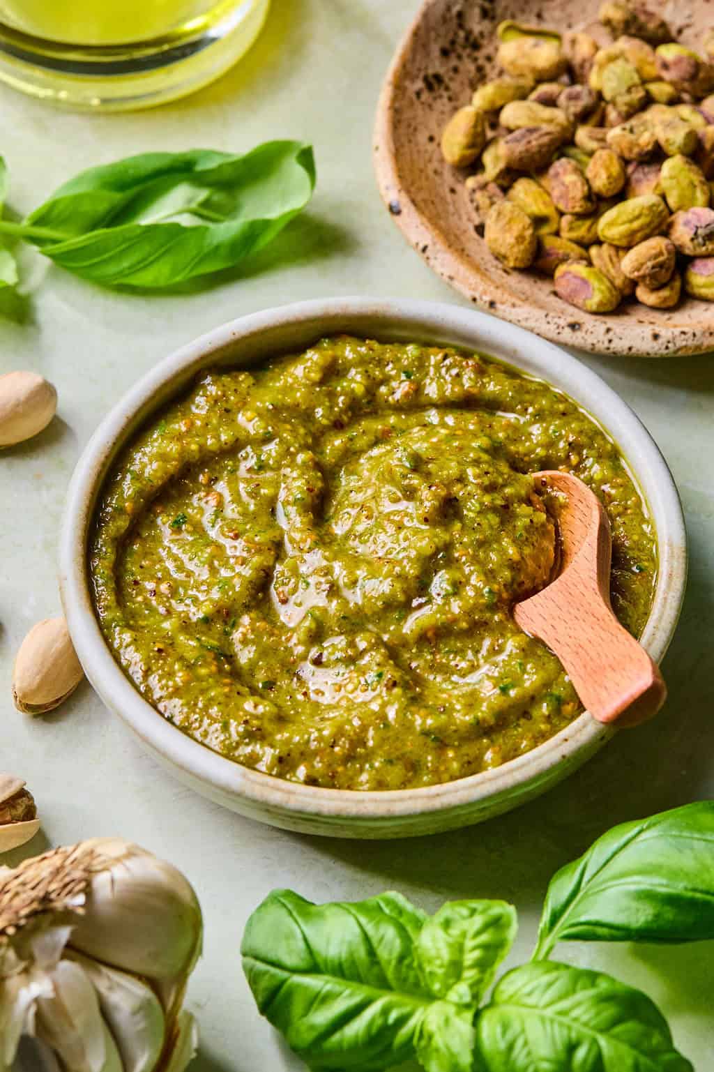 A bowl of green pistachio pesto with a small wooden spoon, surrounded by fresh basil leaves, garlic, and a dish of shelled pistachios on a light surface.