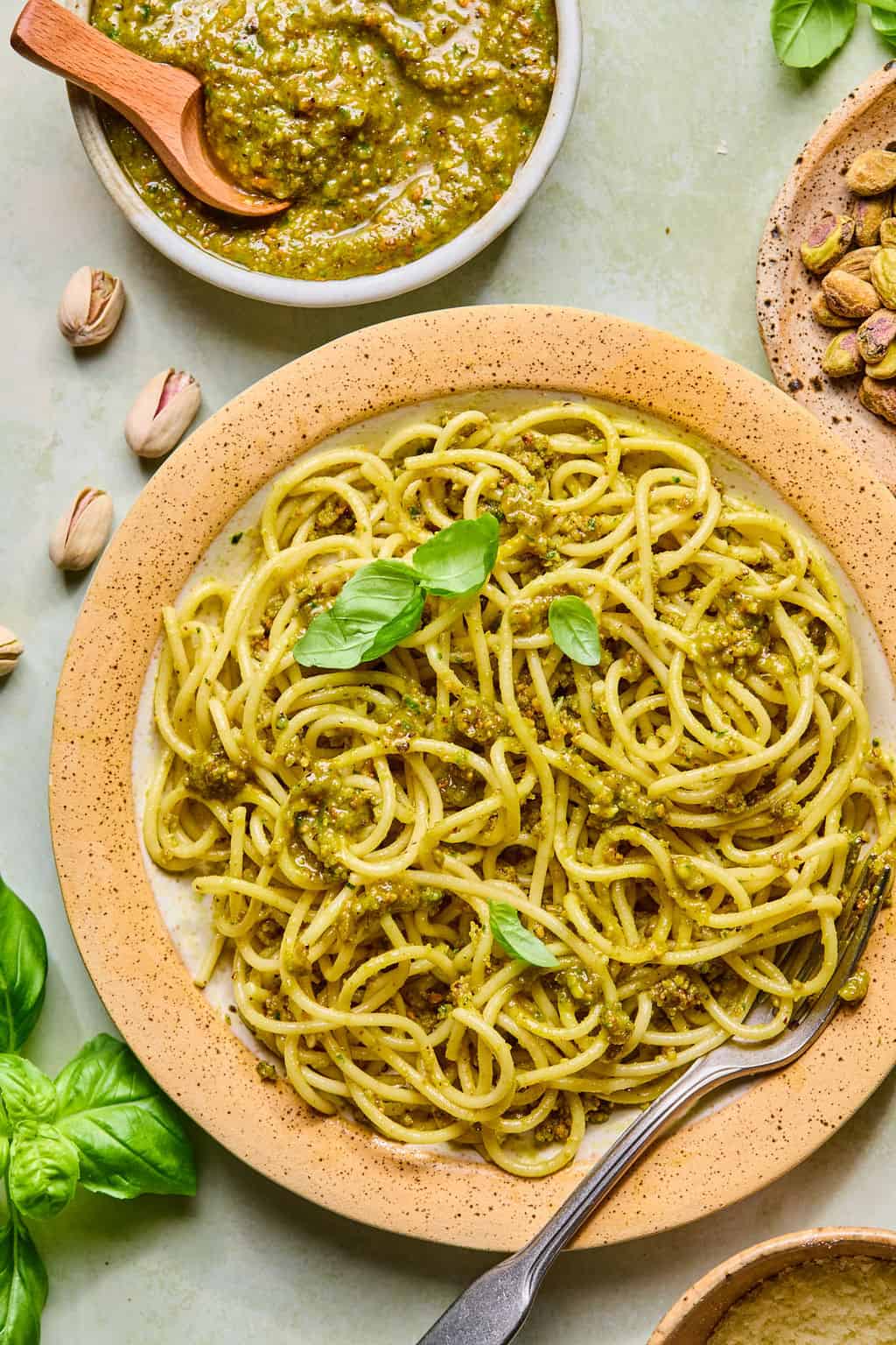 A plate of spaghetti topped with green pistachio pesto and fresh basil leaves, with a fork on the side. Nearby are a bowl of pesto, scattered pistachios, and basil leaves on a light surface.