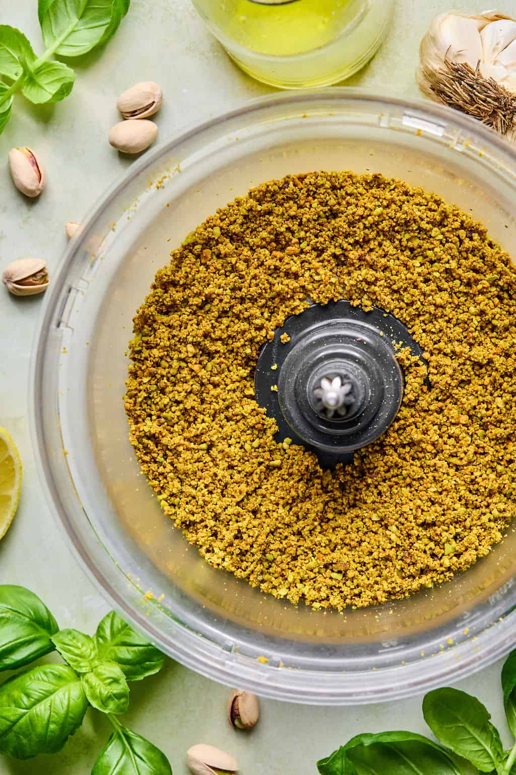A food processor bowl filled with finely ground pistachios, surrounded by fresh basil leaves, whole pistachios, a lemon wedge, garlic, and a bottle of olive oil on a light-colored surface.