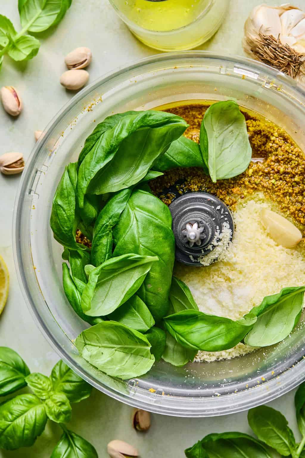 A food processor bowl filled with fresh basil leaves, grated cheese, garlic, pistachios, and olive oil, ready to be blended into pesto. Surrounding the bowl are more basil leaves, pistachios, garlic, and a lemon slice.