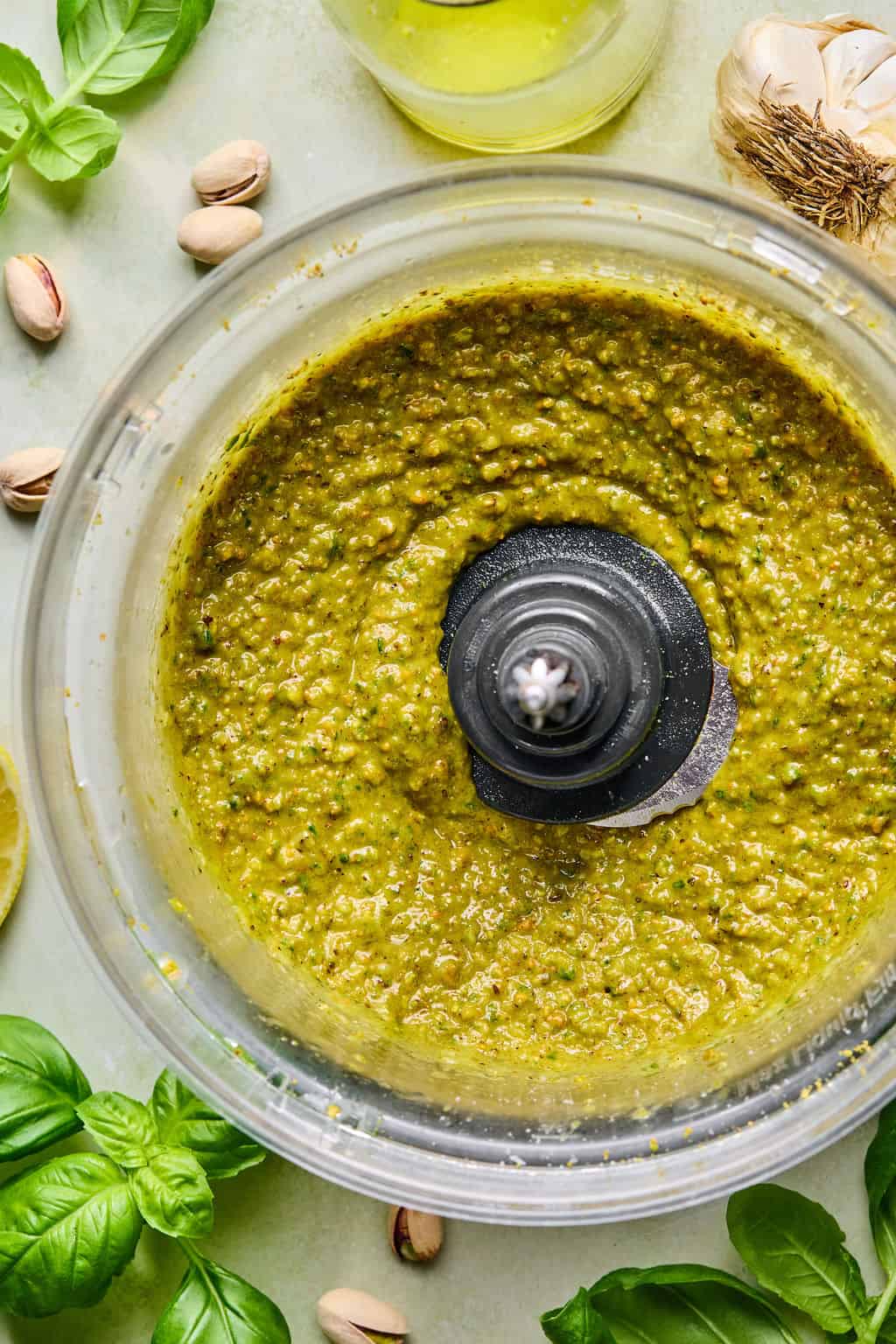 A food processor bowl filled with freshly blended green pistachio pesto, surrounded by basil leaves, garlic, pistachios, lemon, and a bottle of olive oil on a light surface.