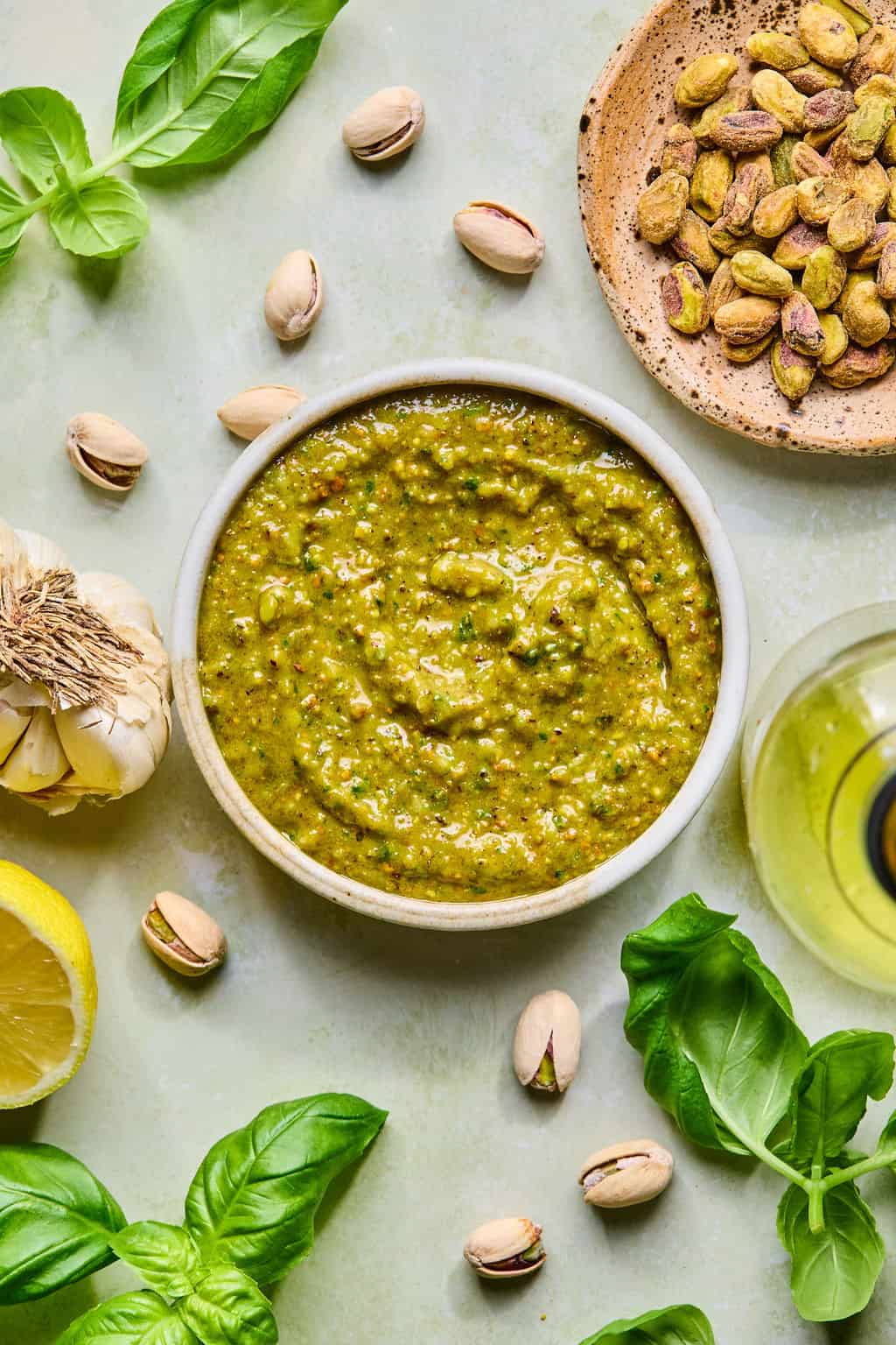 A bowl of green pistachio pesto surrounded by fresh basil leaves, whole pistachios, garlic, a half lemon, a bottle of olive oil, and a small plate of shelled pistachios on a light surface.