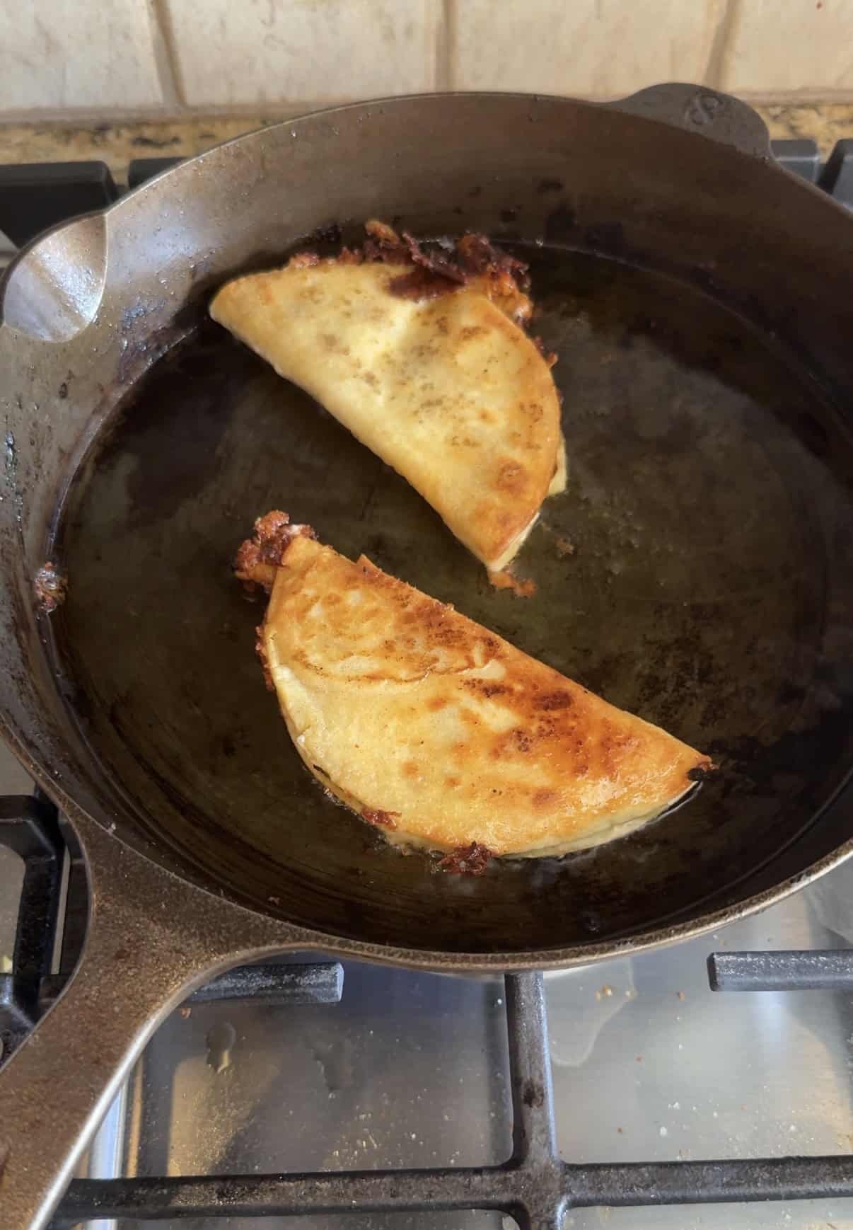 Two golden-brown crispy chicken tacos with melted cheese and filling are cooking in a black cast iron skillet on a stovetop. The tacos are folded in half and appear crispy around the edges.