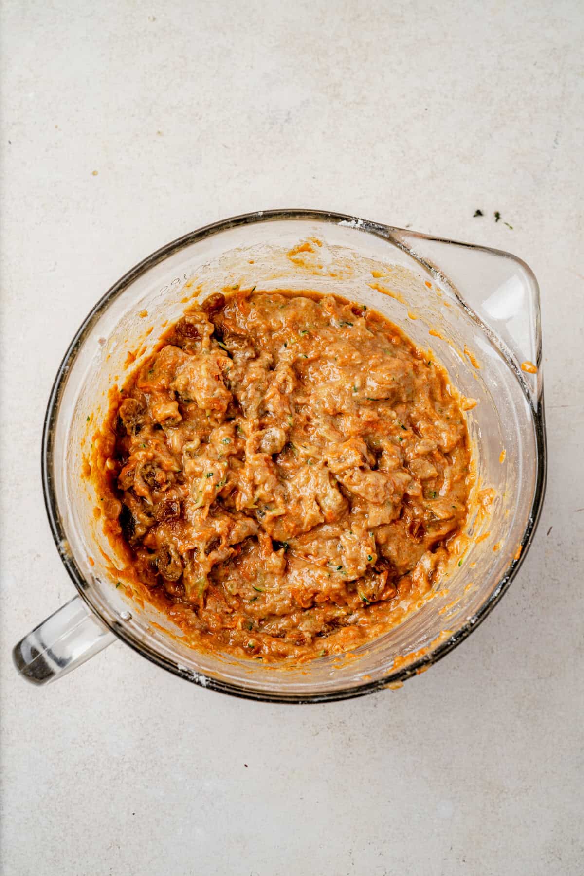 A glass mixing bowl filled with a thick zucchini carrot muffin batter on a light-colored surface, seen from above.
