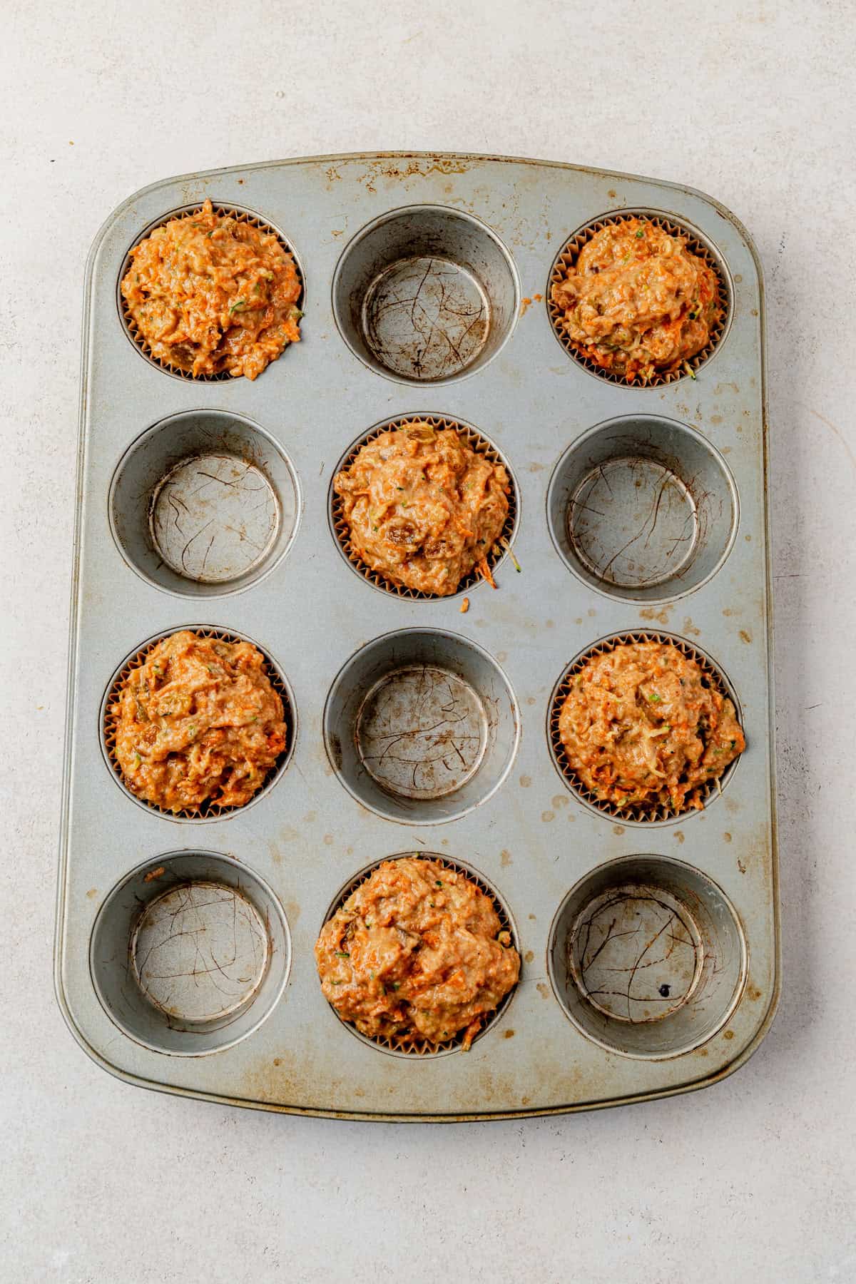 A muffin tin holds five unbaked muffins in paper liners, arranged unevenly among the nine spaces, on a light-colored surface.