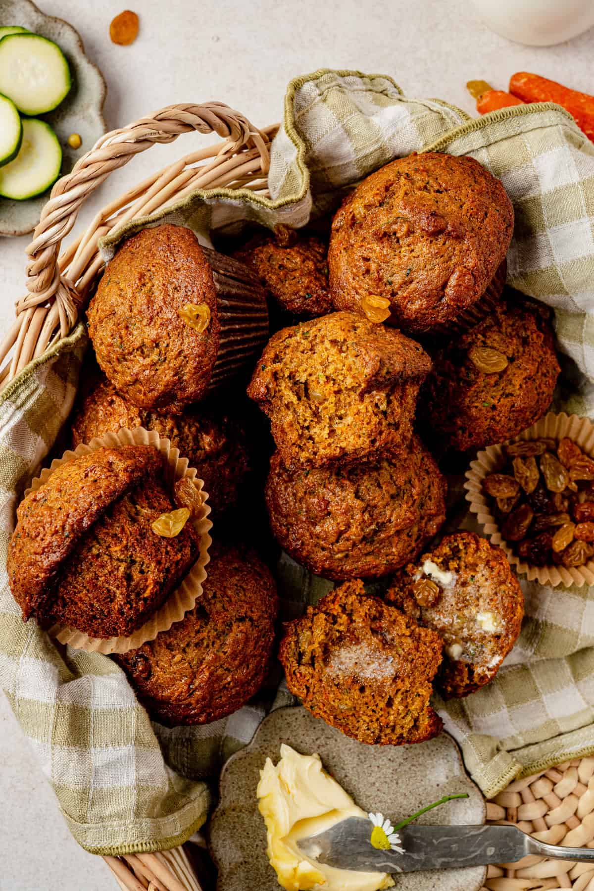 A wicker basket lined with a green checkered cloth holds several zucchini muffins. One muffin is split open with butter melting inside. A butter knife and a slab of butter lie nearby.