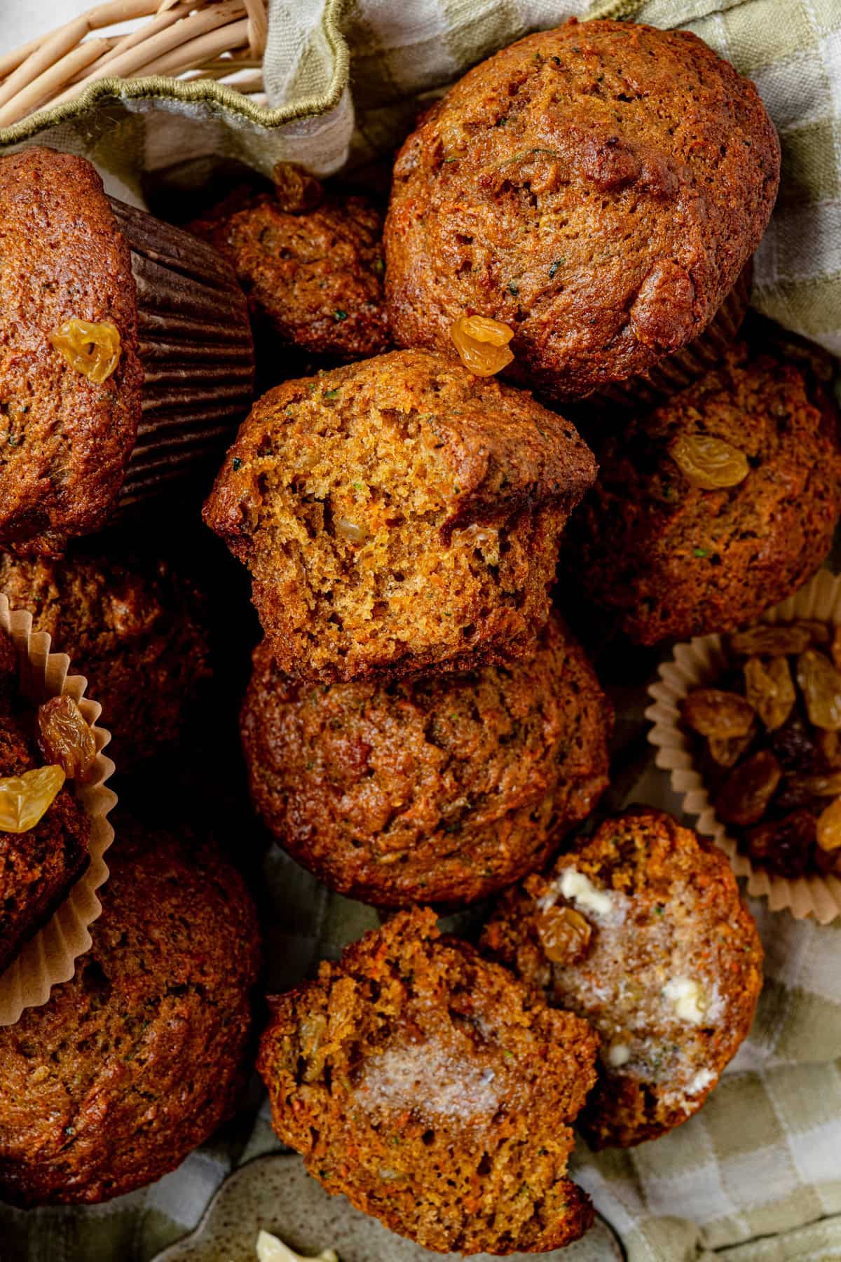 A close-up of freshly baked muffins, some broken open to reveal a moist crumb. Golden raisins are scattered around, and a checkered cloth lines the background.