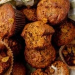 A close-up of freshly baked carrot zucchini muffins with golden raisins, some in paper liners, arranged together. One muffin is broken open, revealing a moist, textured interior.
