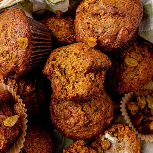 A close-up of freshly baked carrot zucchini muffins with golden raisins, some in paper liners, arranged together. One muffin is broken open, revealing a moist, textured interior.