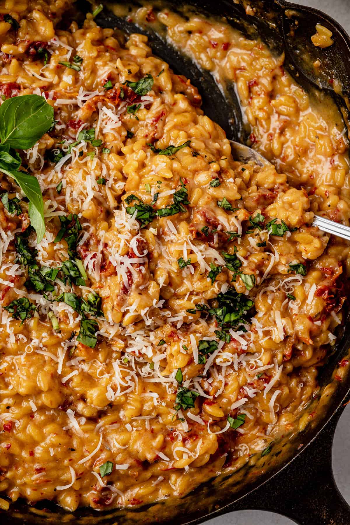 Creamy sun-dried tomato orzo with grated parmesan cheese, and fresh chopped basil, served in a cast-iron skillet with a spoon.