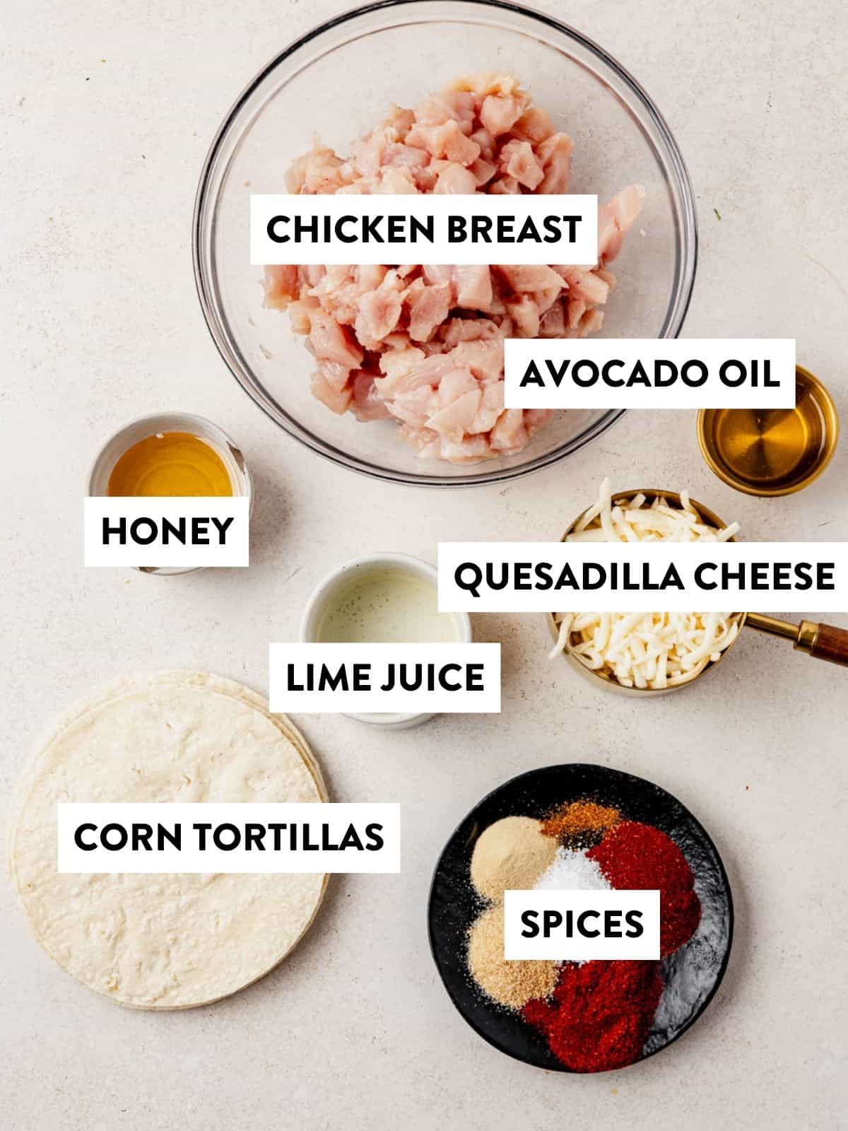 A top-down view of labeled ingredients on a white surface: diced chicken breast, avocado oil, honey, quesadilla cheese, lime juice, corn tortillas, and a small plate of assorted spices.