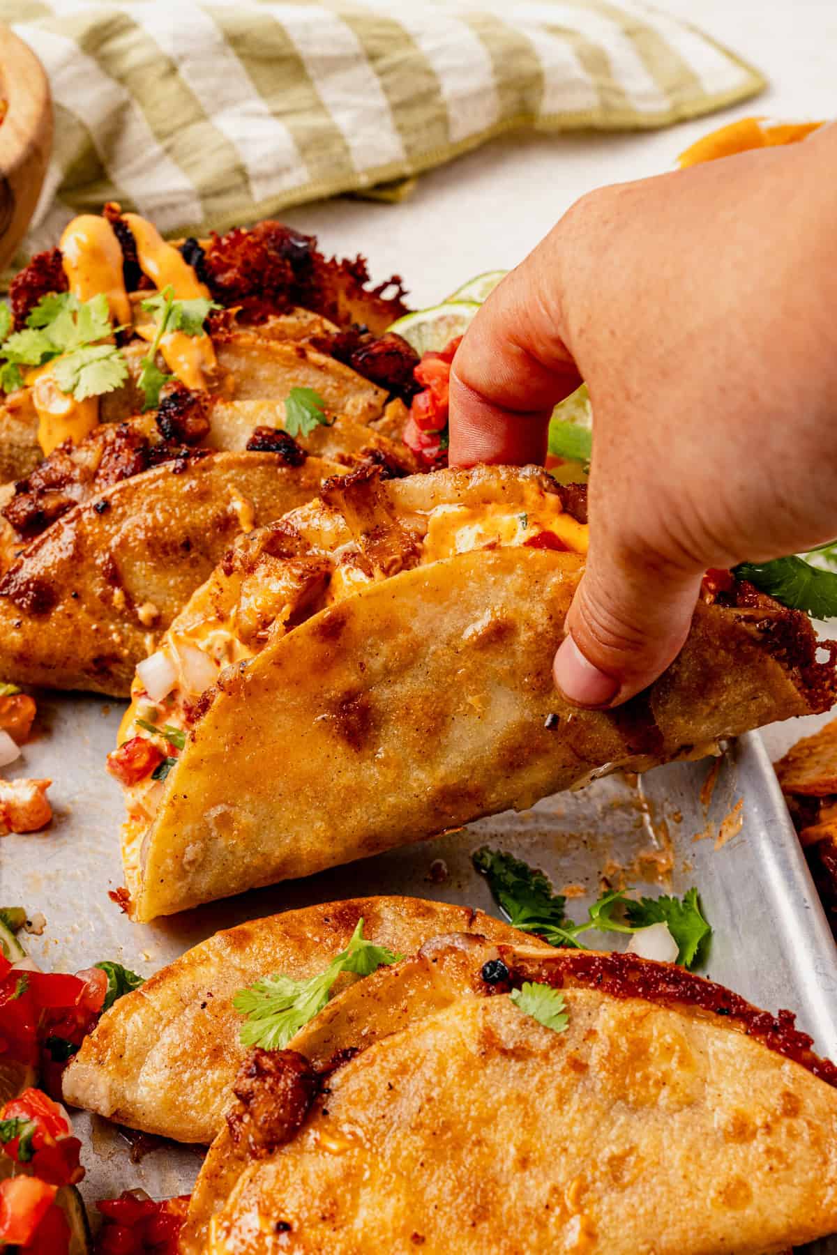 A hand is picking up a crispy, golden-brown crispy chicken taco filled with meat, cheese, and fresh toppings from a tray of tacos, with garnishes and a green-striped napkin in the background.