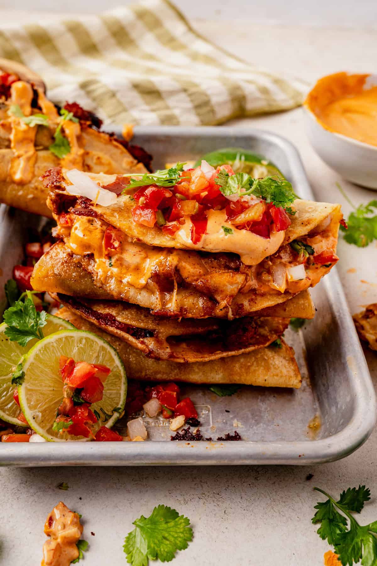 A metal tray with stacked crispy chicken tacos filled with meat, topped with diced tomatoes, onions, cilantro, and creamy orange sauce. Garnished with lime slices and fresh cilantro, with a small bowl of sauce in the background.