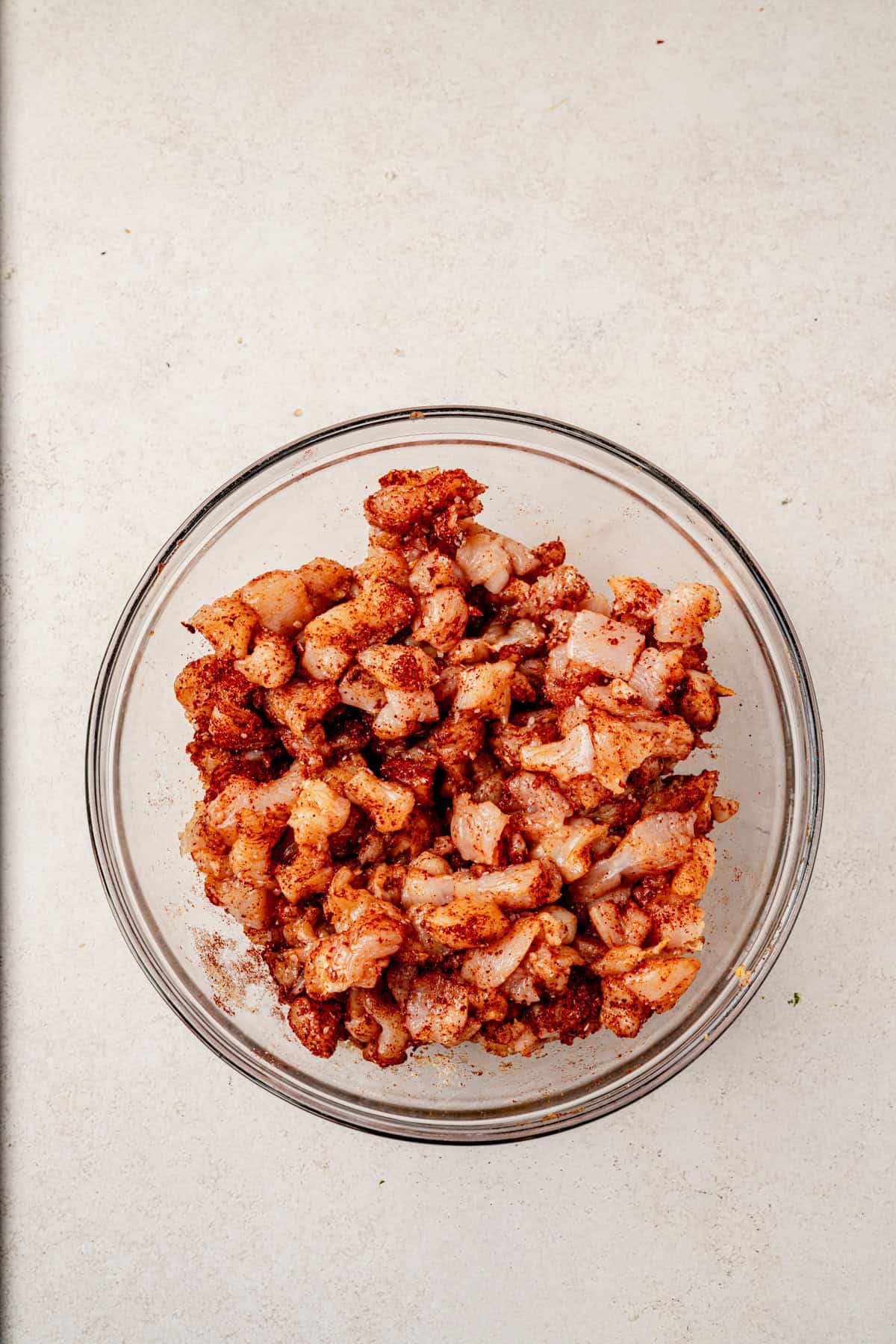 A glass bowl filled with small pieces of raw chicken coated in red spices, set on a light-colored countertop.