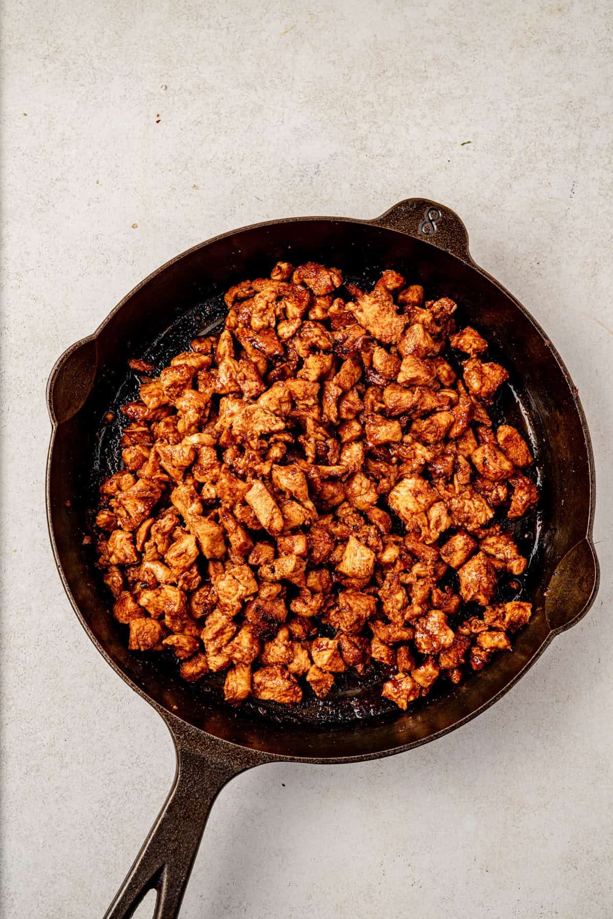 A cast iron skillet filled with cooked, seasoned, and diced chicken pieces sits on a light-colored surface.
