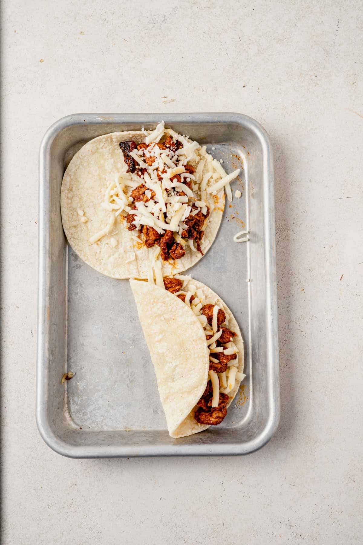 Two corn tortillas filled with seasoned meat and topped with shredded cheese are placed in a rectangular metal baking pan on a light-colored surface. One taco is folded while the other is open-faced.