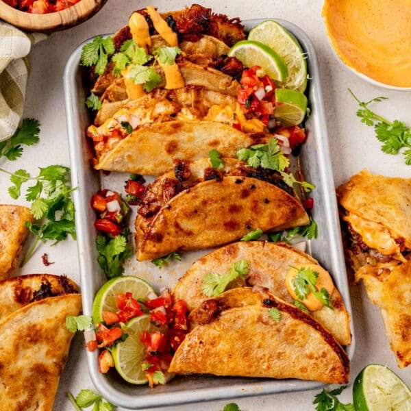 A tray of cheesy, crispy chicken tacos filled with meat, topped with fresh cilantro, tomato salsa, and lime wedges, surrounded by more tacos, cilantro leaves, and a bowl of sauce on a light surface.