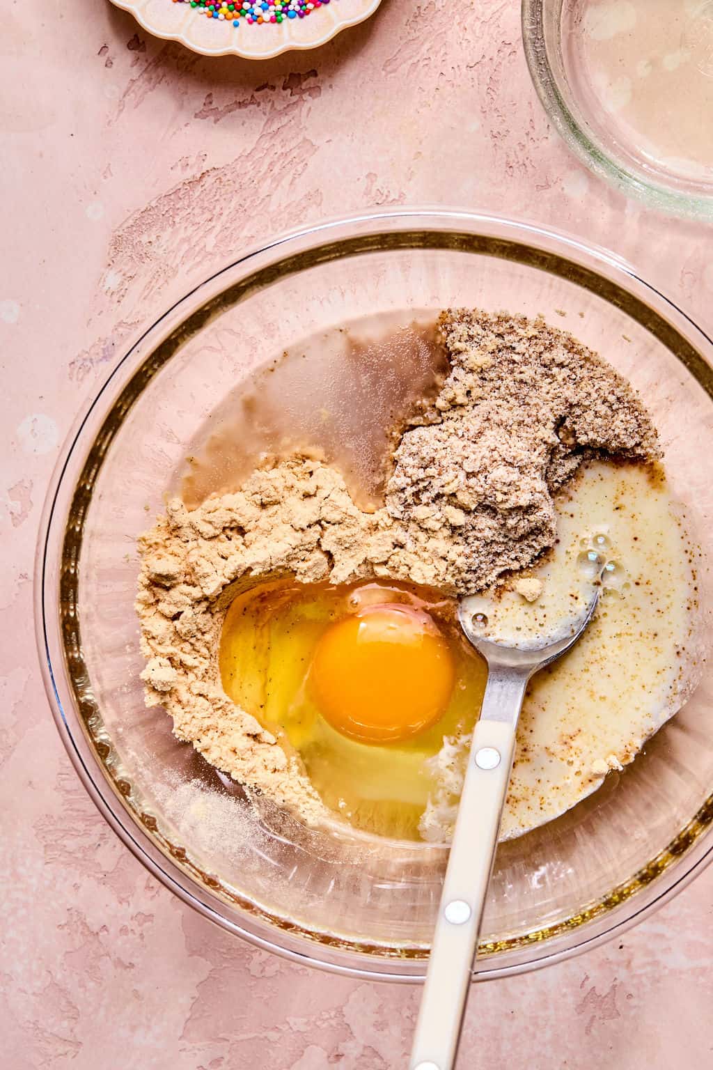 A glass bowl contains flour, ground nuts, an egg, and liquid, partially mixed with a white-handled fork. Sprinkles and another glass bowl are visible nearby on a pink textured surface.