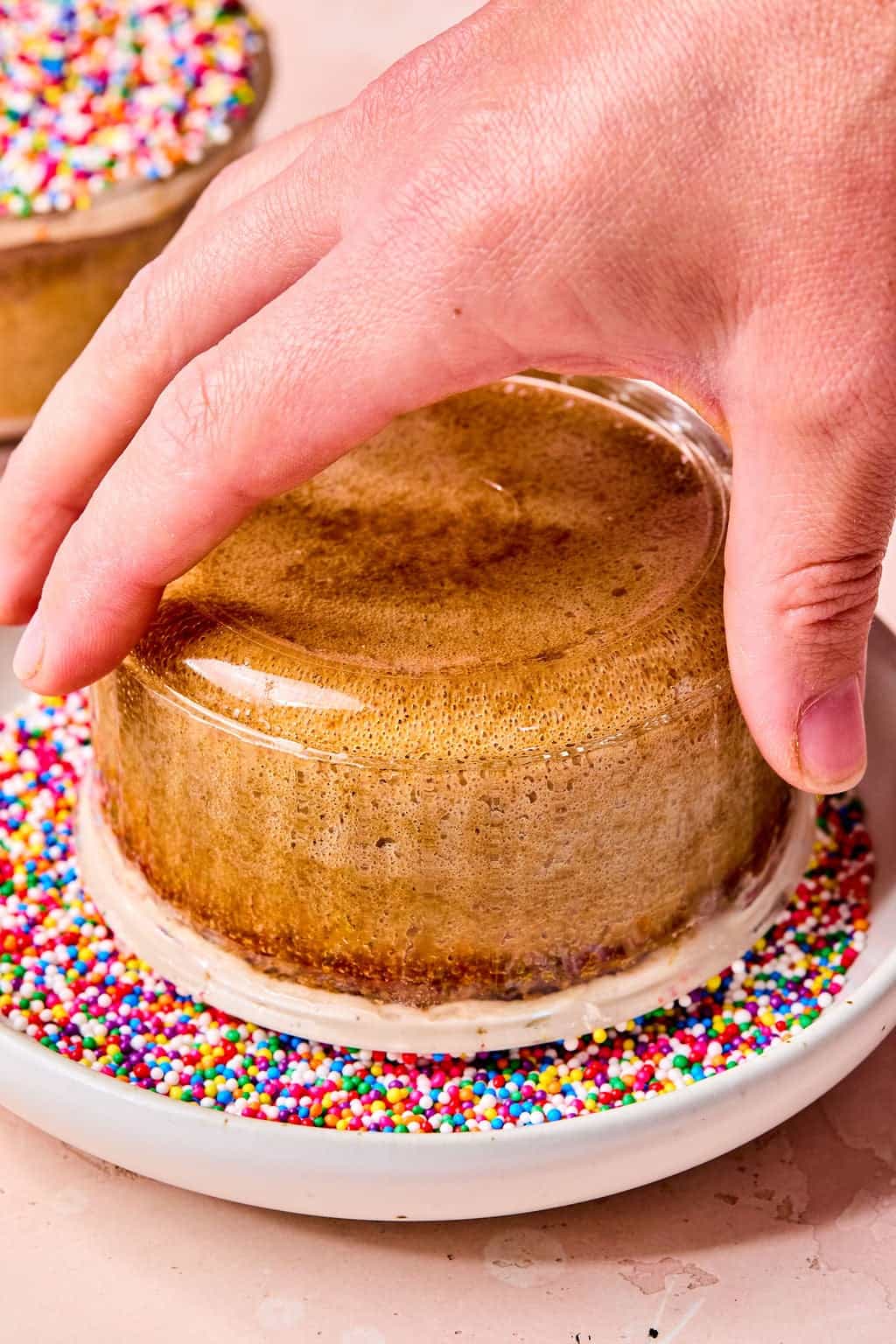 A hand lifts a small round cake, still in its glass dish, and dips the rim in colorful rainbow sprinkles on a white plate.