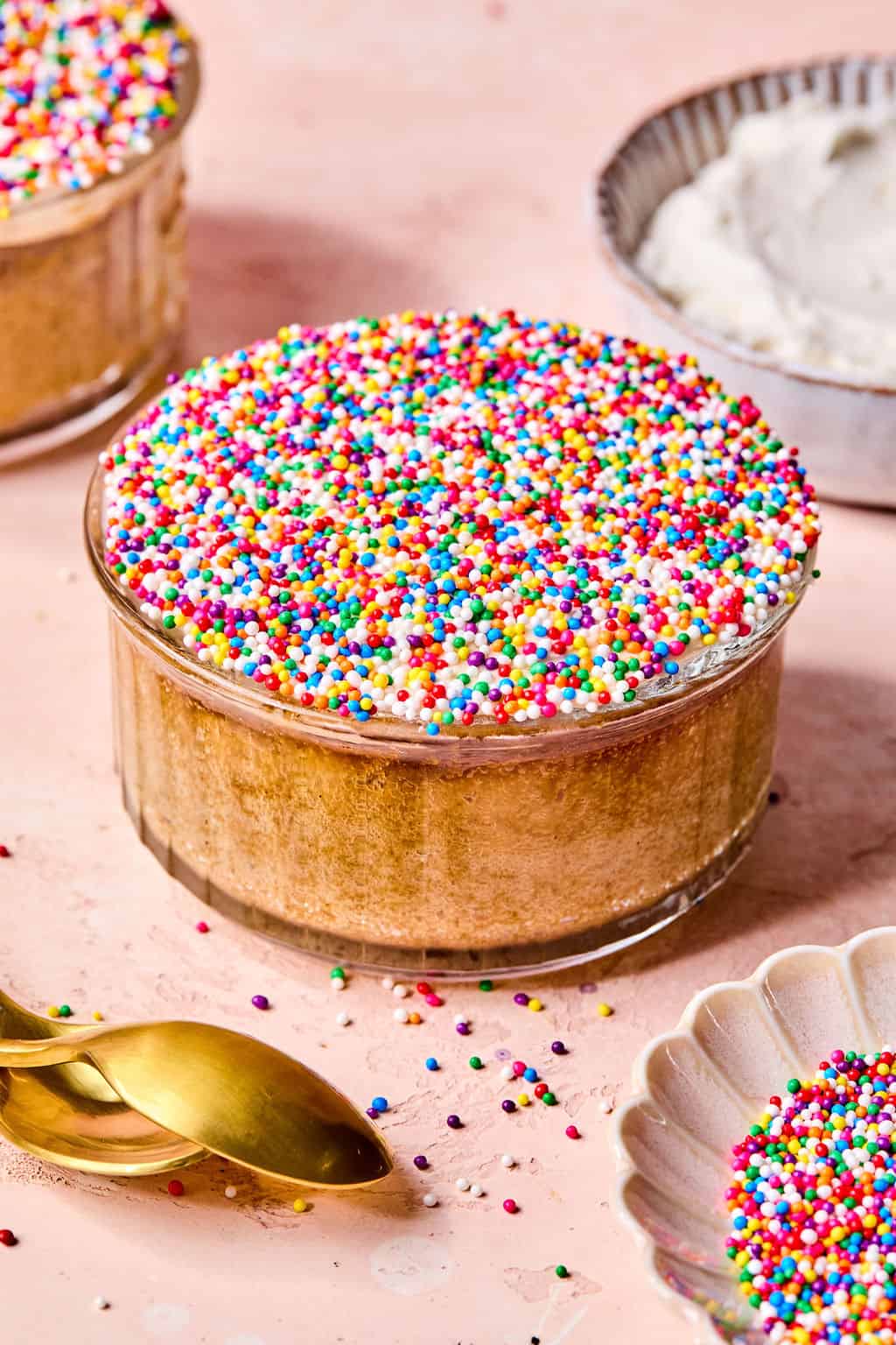 A small glass dish filled with cake topped with colorful round sprinkles sits on a pink surface near a spoon and a bowl of white frosting.