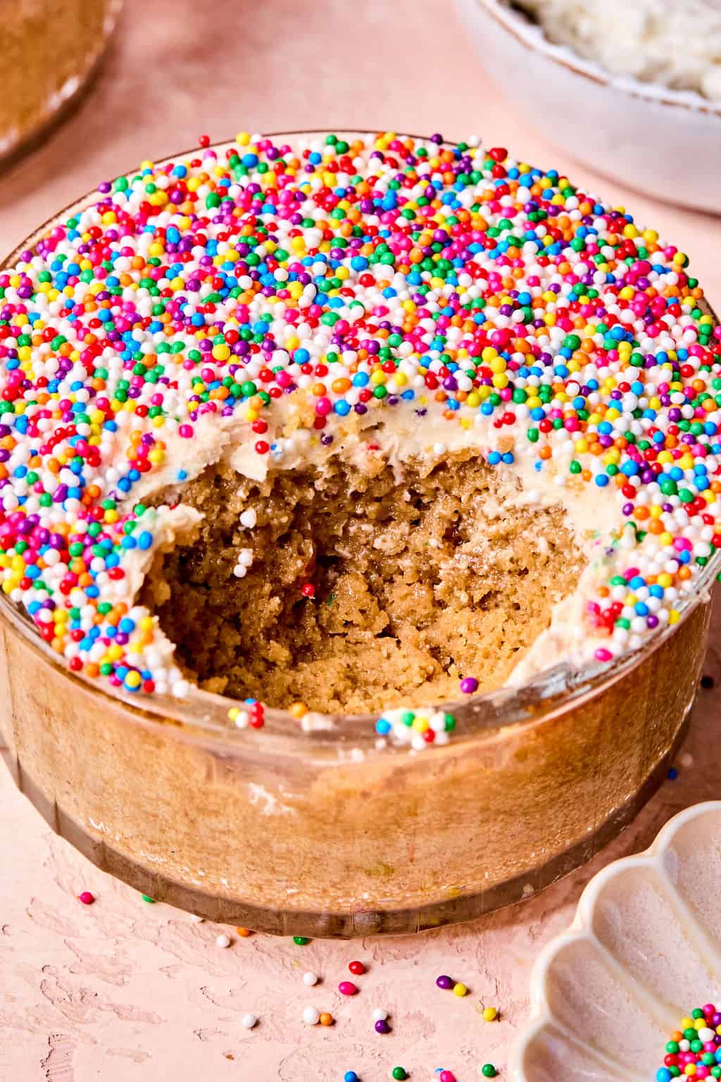 A round glass dish with a partially eaten protein dot cake topped with vanilla protein frosting and colorful round sprinkles, sitting on a light pink surface.