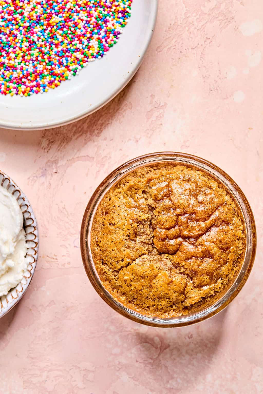 A single-serve baked cake in a glass jar sits on a pink surface, with a bowl of rainbow sprinkles and a bowl of white frosting nearby.