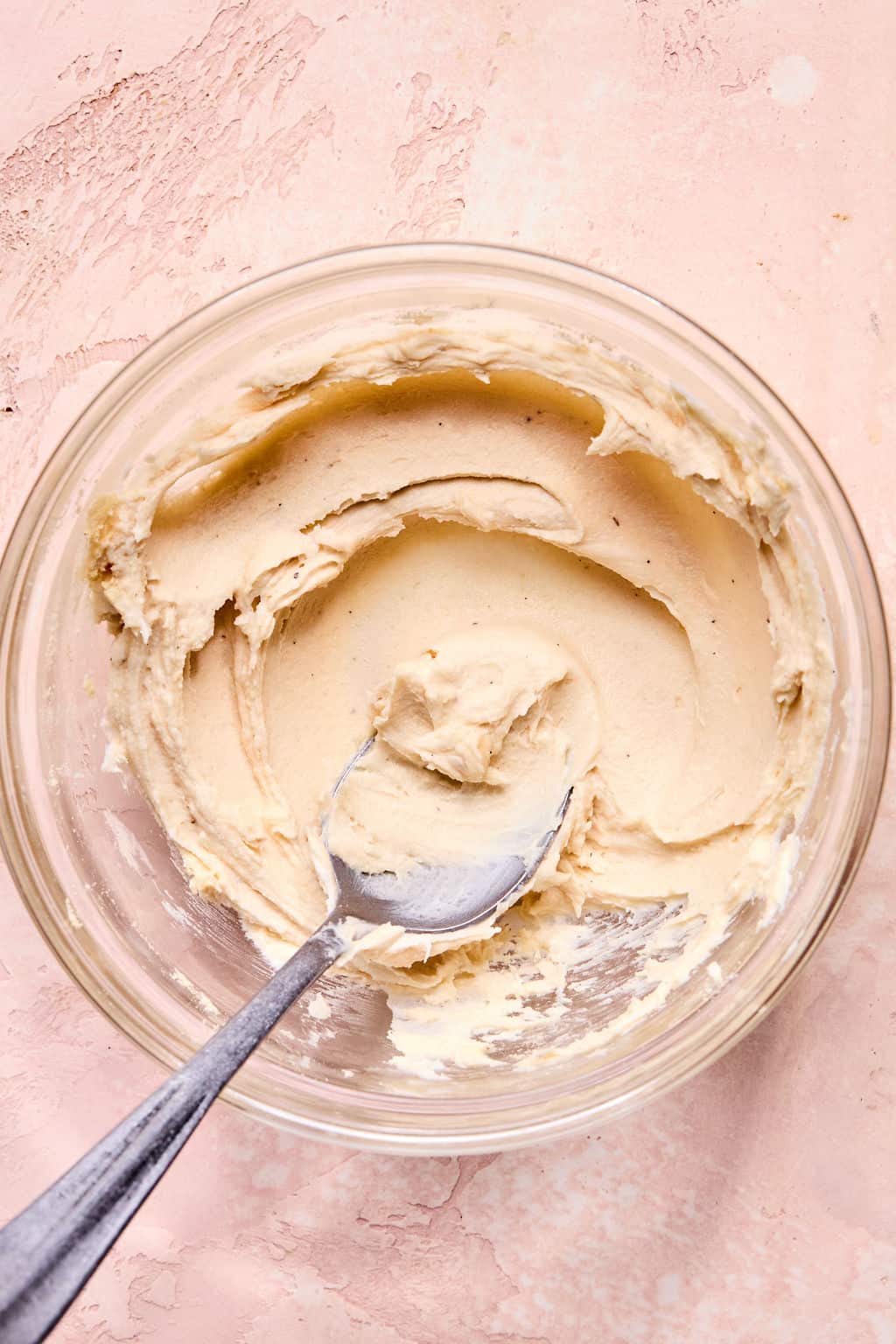 A glass bowl with creamy, light tan cookie dough and a metal spoon resting inside, set on a pink textured surface.