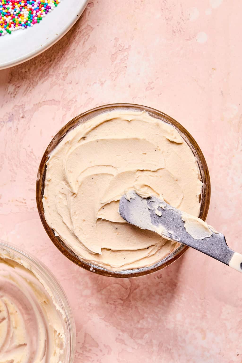 A bowl of light brown frosting with a spatula resting on top, set on a pink surface. A plate of colorful sprinkles is partially visible in the corner.
