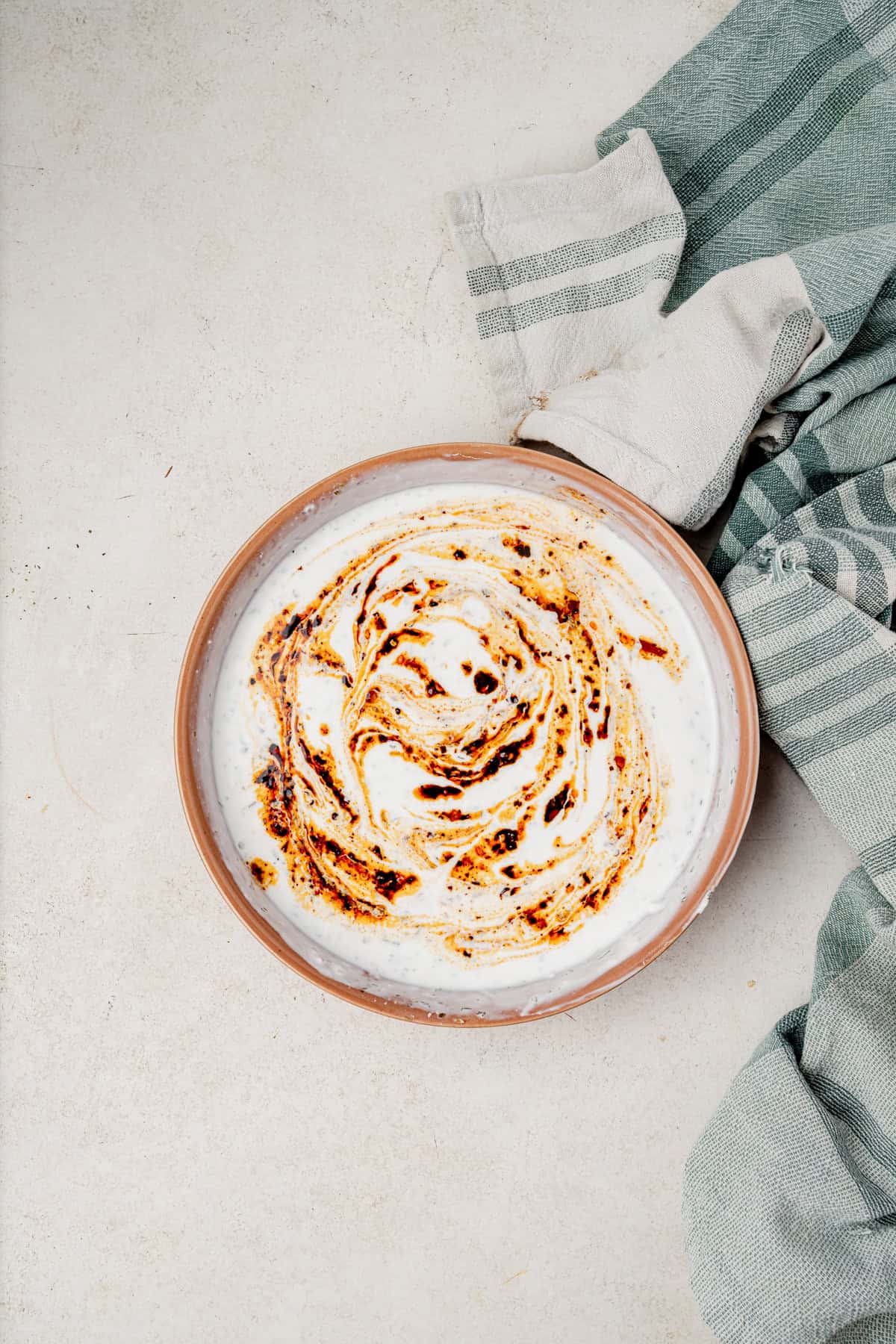 A bowl of yogurt dressing topped with a swirl of chili oil sits on a light surface, next to two folded green and white striped kitchen towels.