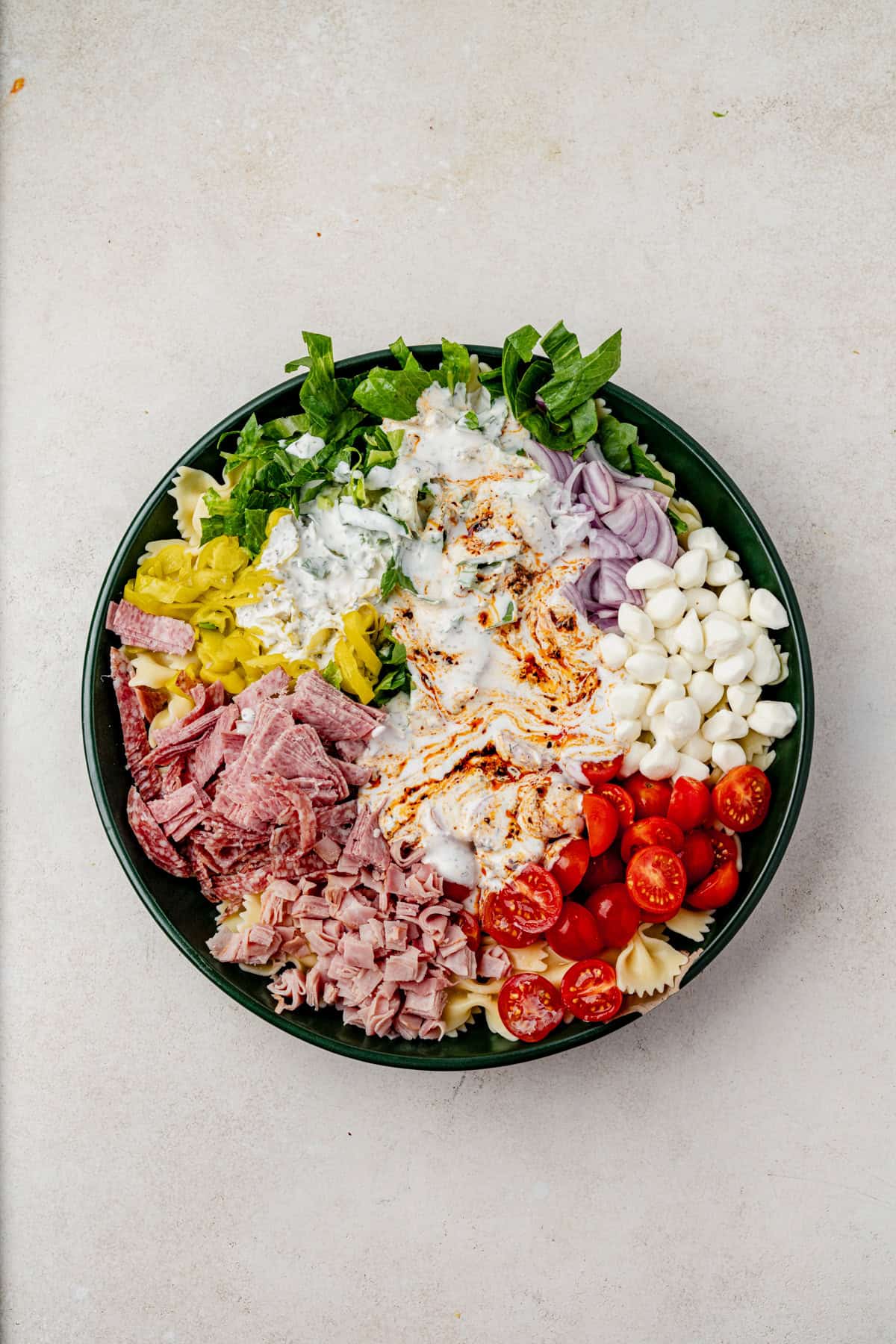A bowl of grinder pasta salad filled with chopped lettuce, sliced onions, mozzarella balls, cherry tomatoes, deli meats, pepperoncini, and pasta, topped with creamy dressing and chili crisp.