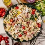 A large bowl of bowtie grinder pasta salad with tomatoes, lettuce, onions, pepperoncini, ham, and creamy dressing, surrounded by salad ingredients on a table.