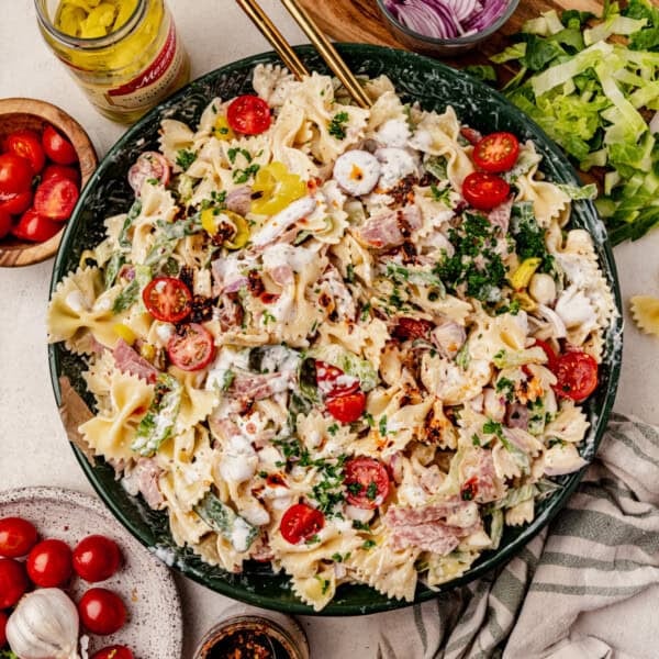 A large bowl of bowtie grinder pasta salad with tomatoes, lettuce, onions, pepperoncini, ham, and creamy dressing, surrounded by salad ingredients on a table.