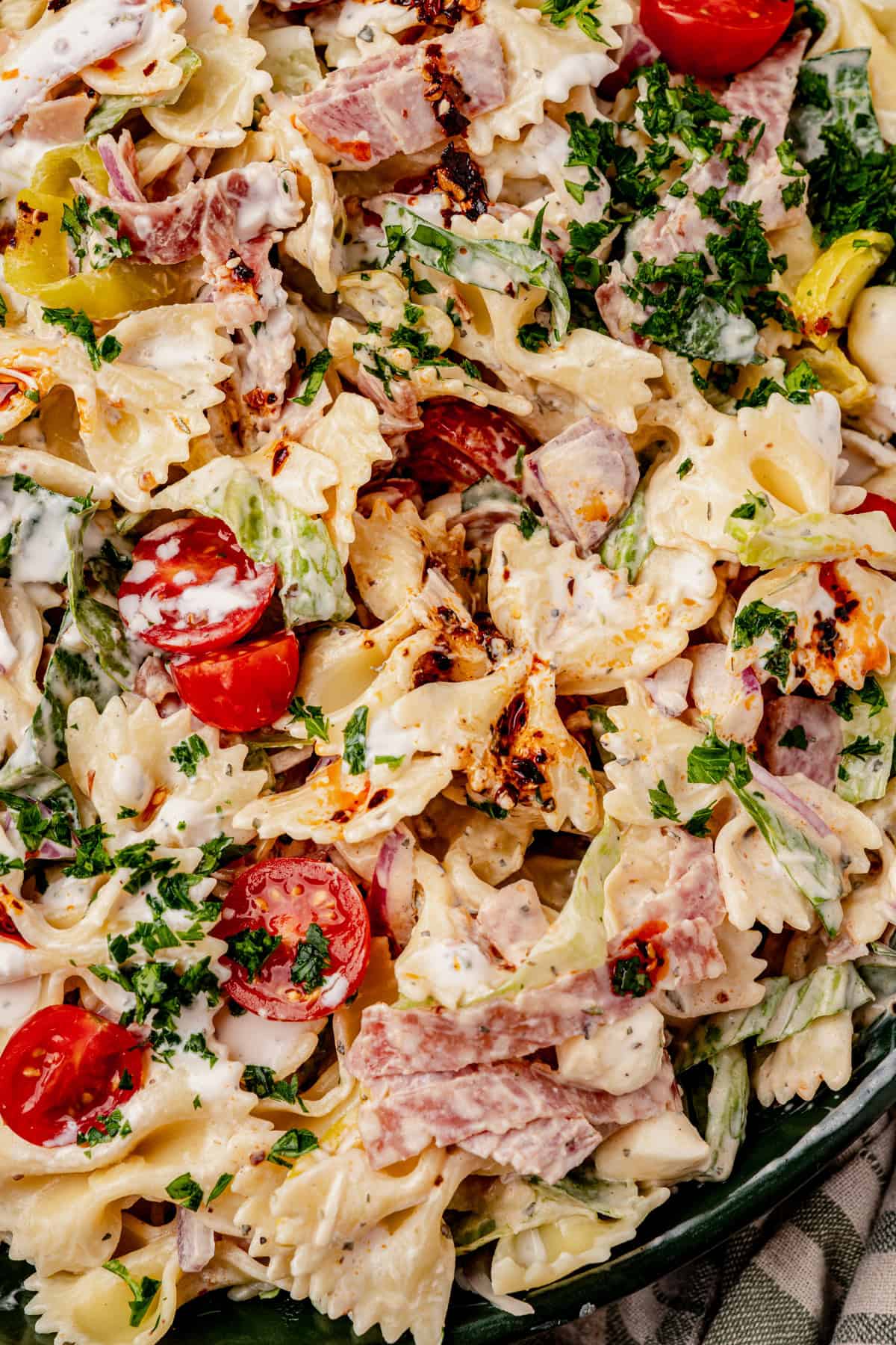 Close-up of a creamy grinder pasta salad with bowtie pasta, cherry tomatoes, lettuce, parsley, ham slices, onions, and a drizzle of dressing, all mixed together.