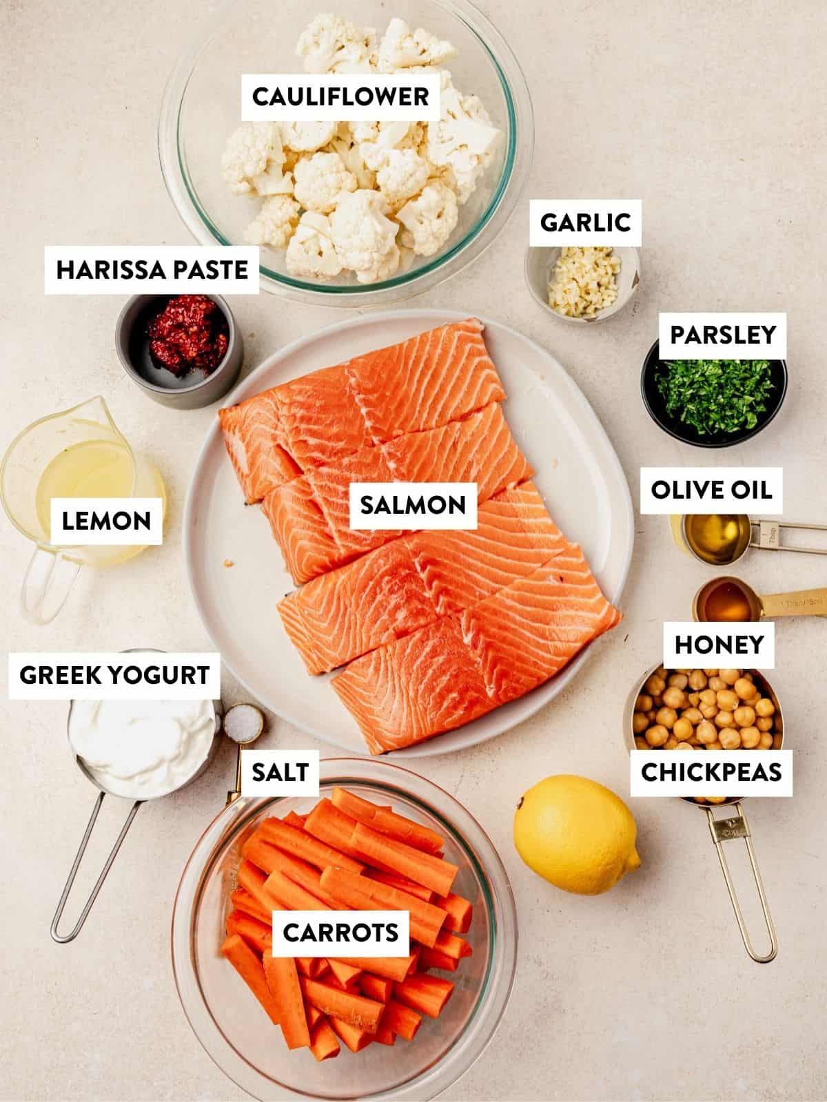 Various labeled ingredients on a countertop, including salmon ready for harissa salmon, cauliflower, garlic, parsley, olive oil, honey, chickpeas, lemon, salt, Greek yogurt, harissa paste, and carrot sticks in bowls and measuring cups.