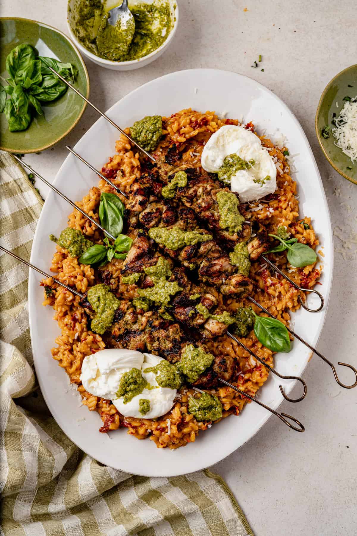 A platter of grilled pesto chicken served over tomato orzo, topped with green pesto sauce and dollops of burrata cheese, garnished with fresh basil leaves. Pesto sauce, basil, and grated cheese are in small bowls nearby.
