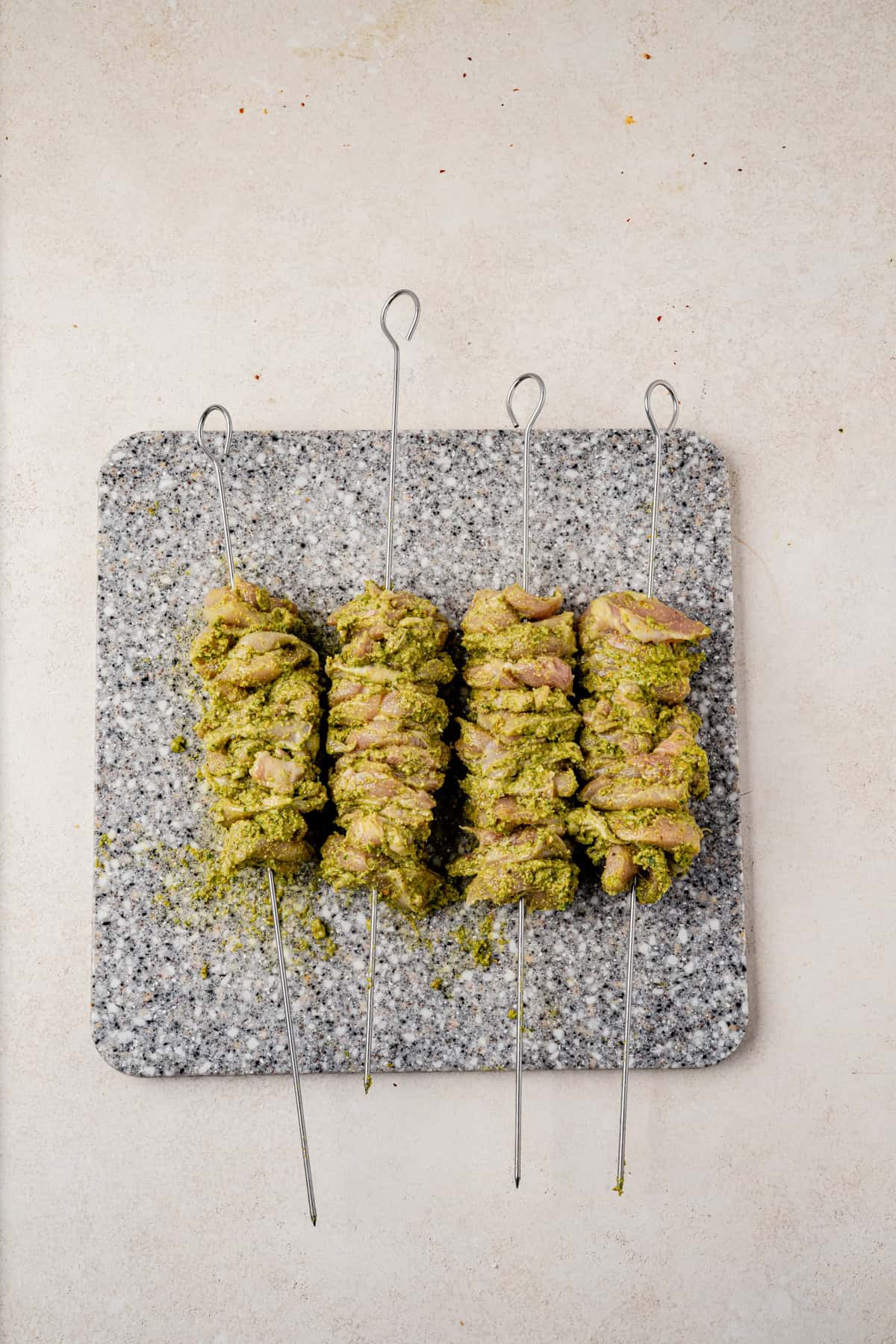 Four metal skewers with pieces of marinated pesto grilled chicken, coated in pesto, are arranged on a rectangular grey speckled cutting board set on a light-colored surface.