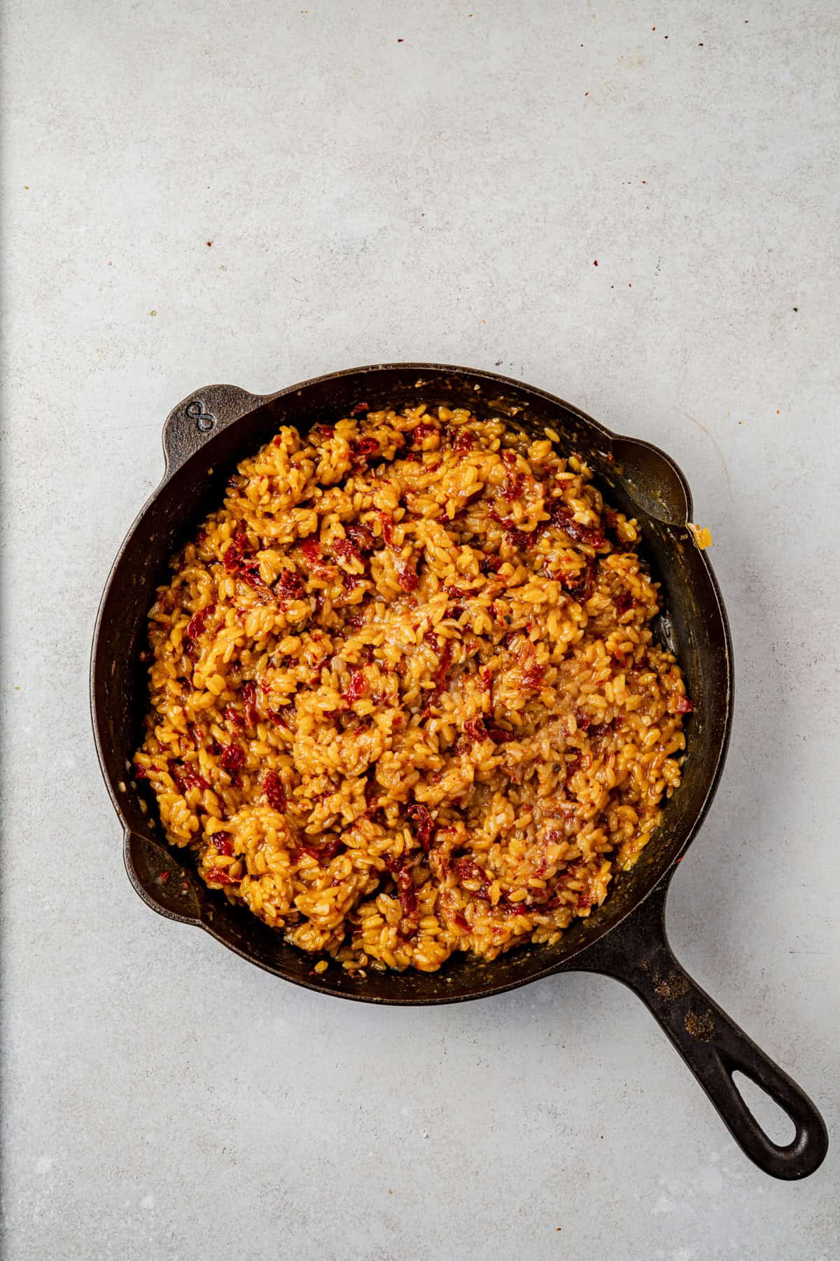 A cast iron skillet filled with creamy sun dried tomato orzo mixed with fresh herbs sits on a light-colored surface.