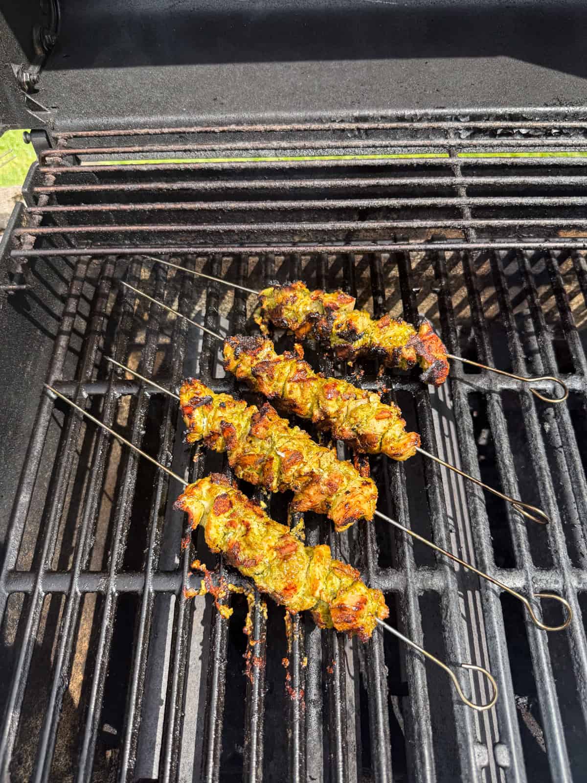 Four metal skewers with marinated pesto chicken are sizzling on an outdoor barbecue grill, with the grill lid open and tasty char marks visible on the food.