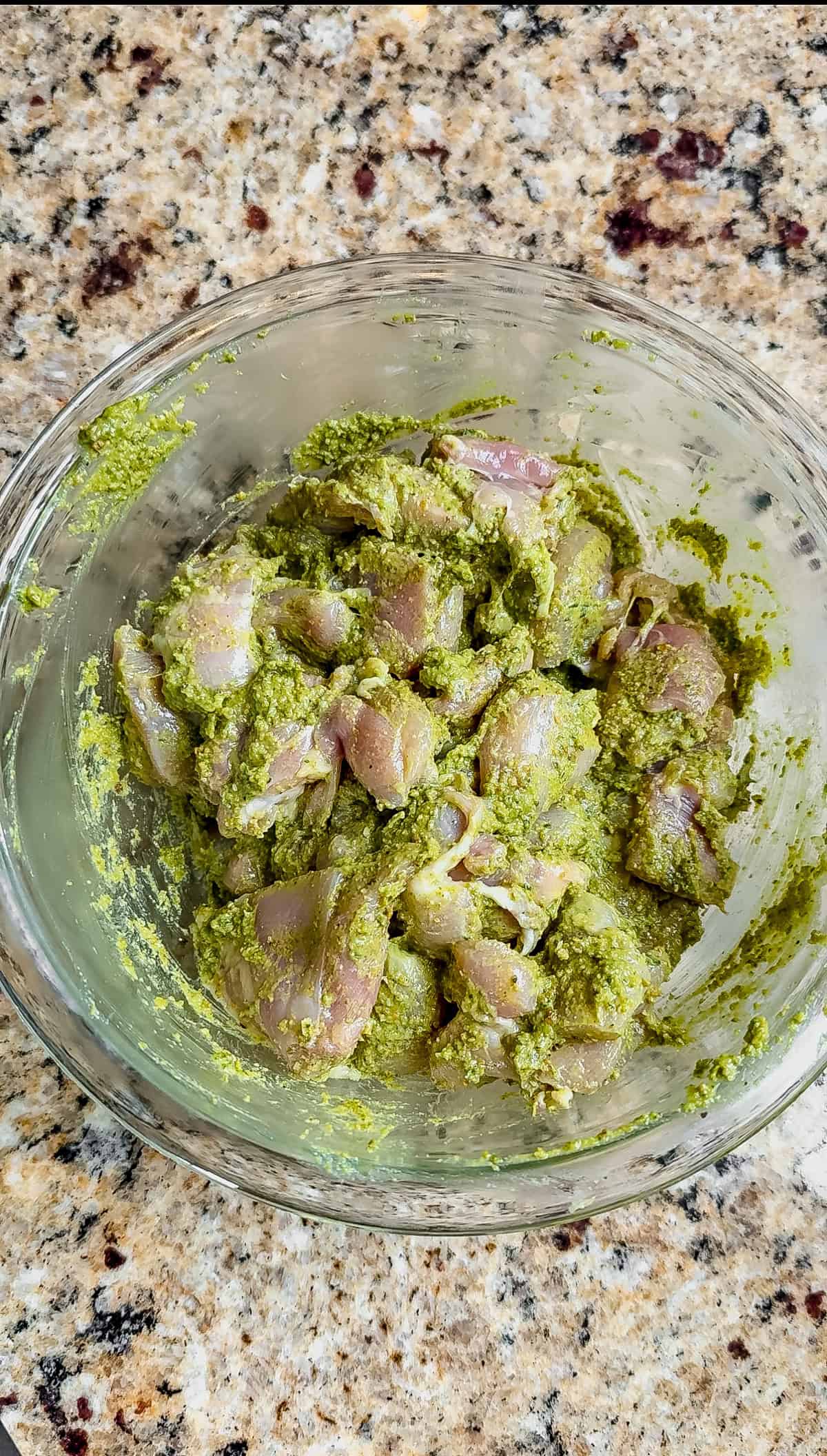 Raw chicken pieces coated in a vibrant green pesto marinade are in a clear glass bowl, sitting on a speckled granite countertop—perfect for making delicious pesto grilled chicken.