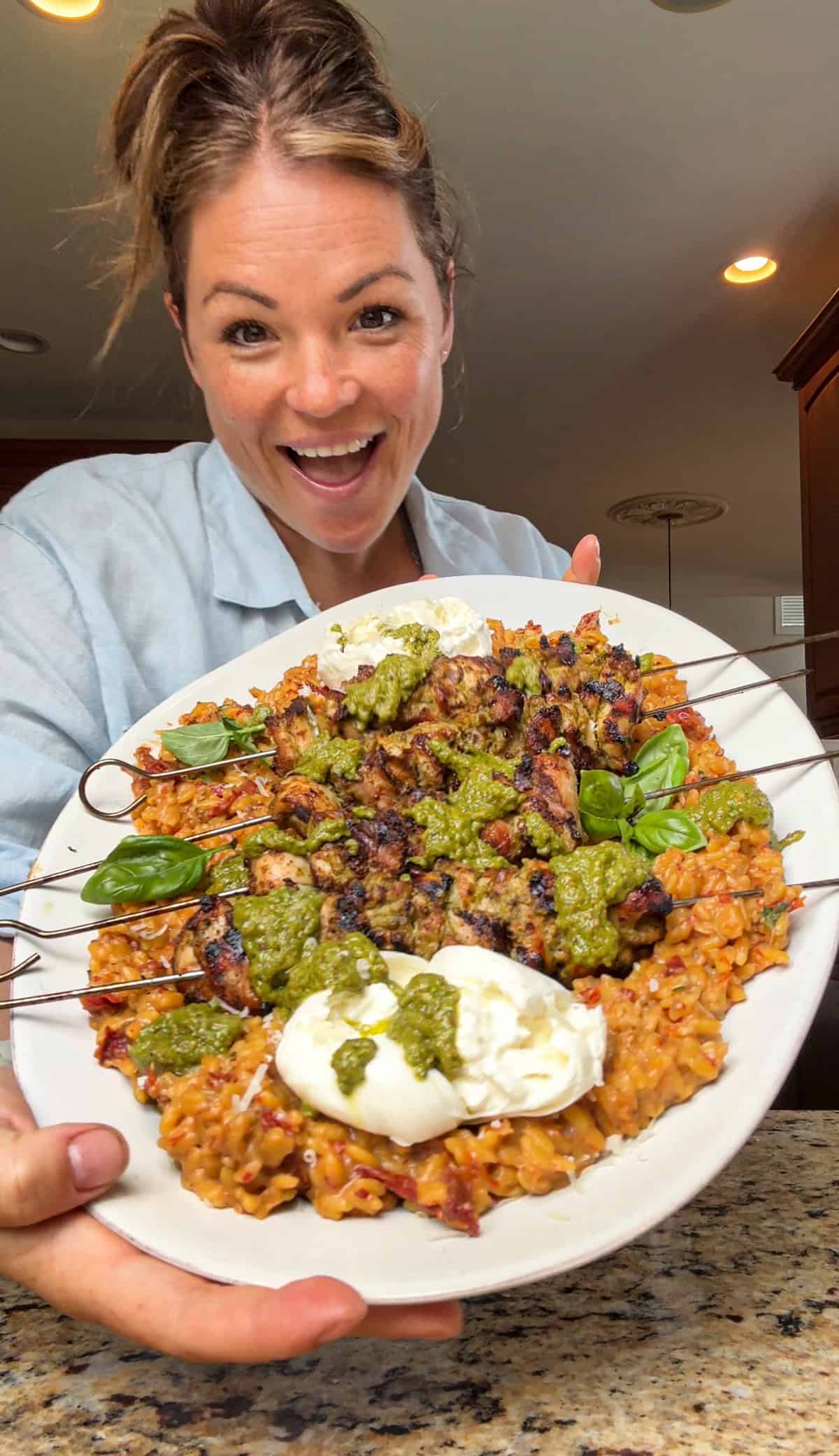 A smiling woman holds a large plate of pesto grilled chicken skewers on a bed of orzo, topped with extra pesto and creamy burrata cheese, garnished with fresh basil.