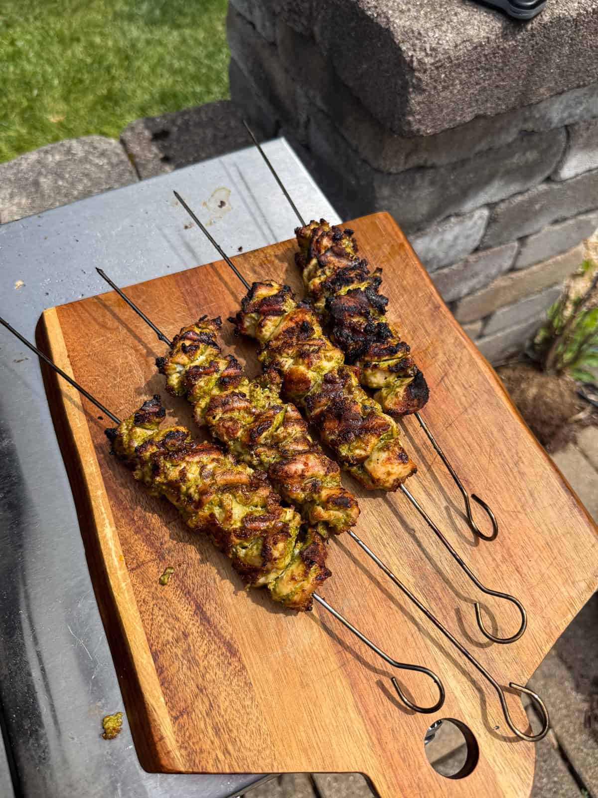 Four pesto grilled chicken skewers with a vibrant green herb marinade rest on a wooden cutting board outdoors, next to a stone wall and grill, on a sunny day.