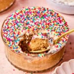 A glass bowl of viral protein dot cake topped with colorful nonpereil sprinkles, with a spoon scooping out a bite, showing the fluffy cake inside.