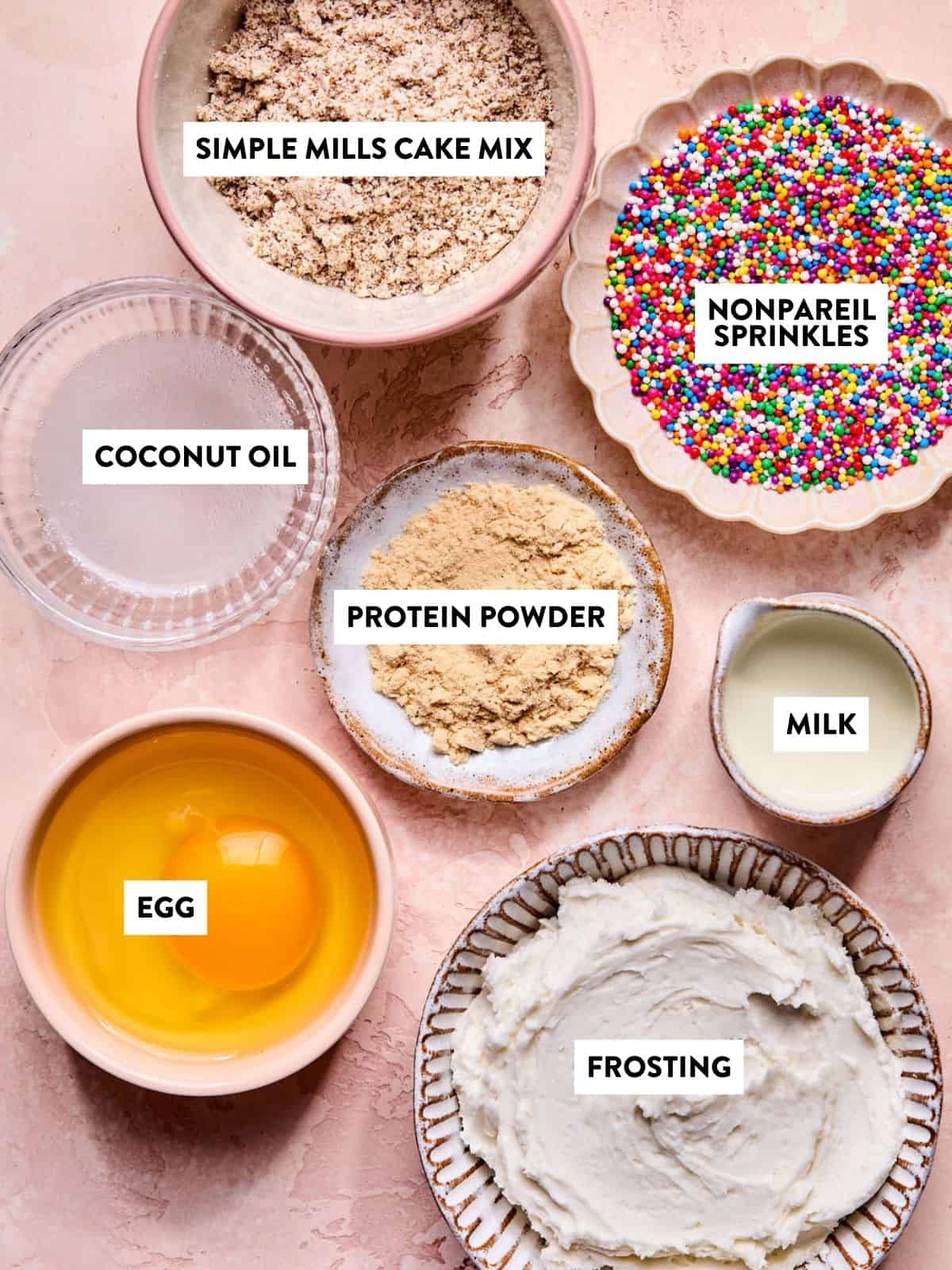 Bowls containing Simple Mills cake mix, nonpareil sprinkles, coconut oil, protein powder, milk, an egg, and frosting are arranged on a pink marbled surface, each labeled with text.