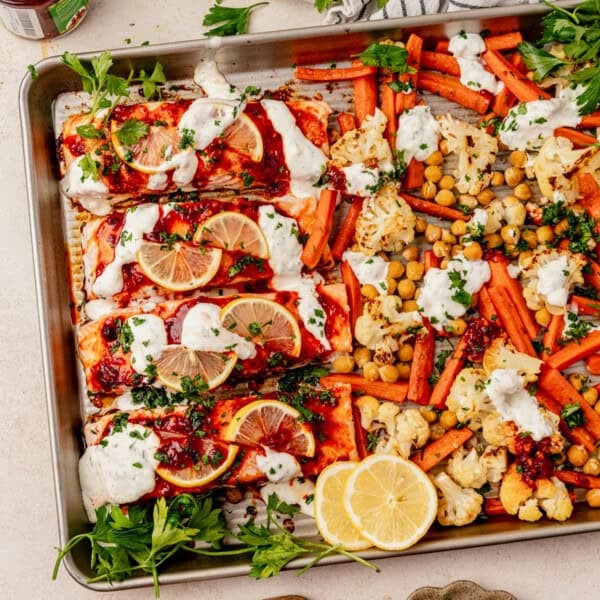 Sheet pan with baked harissa salmon topped with lemon slices and sauce, surrounded by roasted carrots, cauliflower, chickpeas, fresh parsley, and dollops of yogurt sauce. Sliced lemons and herbs garnish the pan.